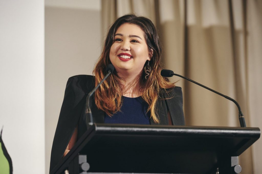 As asian woman standing at a podium with long brown hair smiling to the left of the camera. 