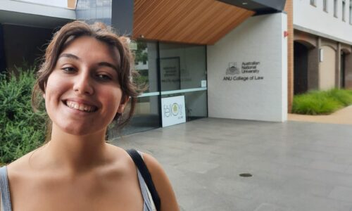 A photo of Margherita smiling in front of the ANU College of Law South Wing building. She has olive skin and brown hair in a bun, and is wearing a grey singlet. The entry to the building and some greenery is behind her.