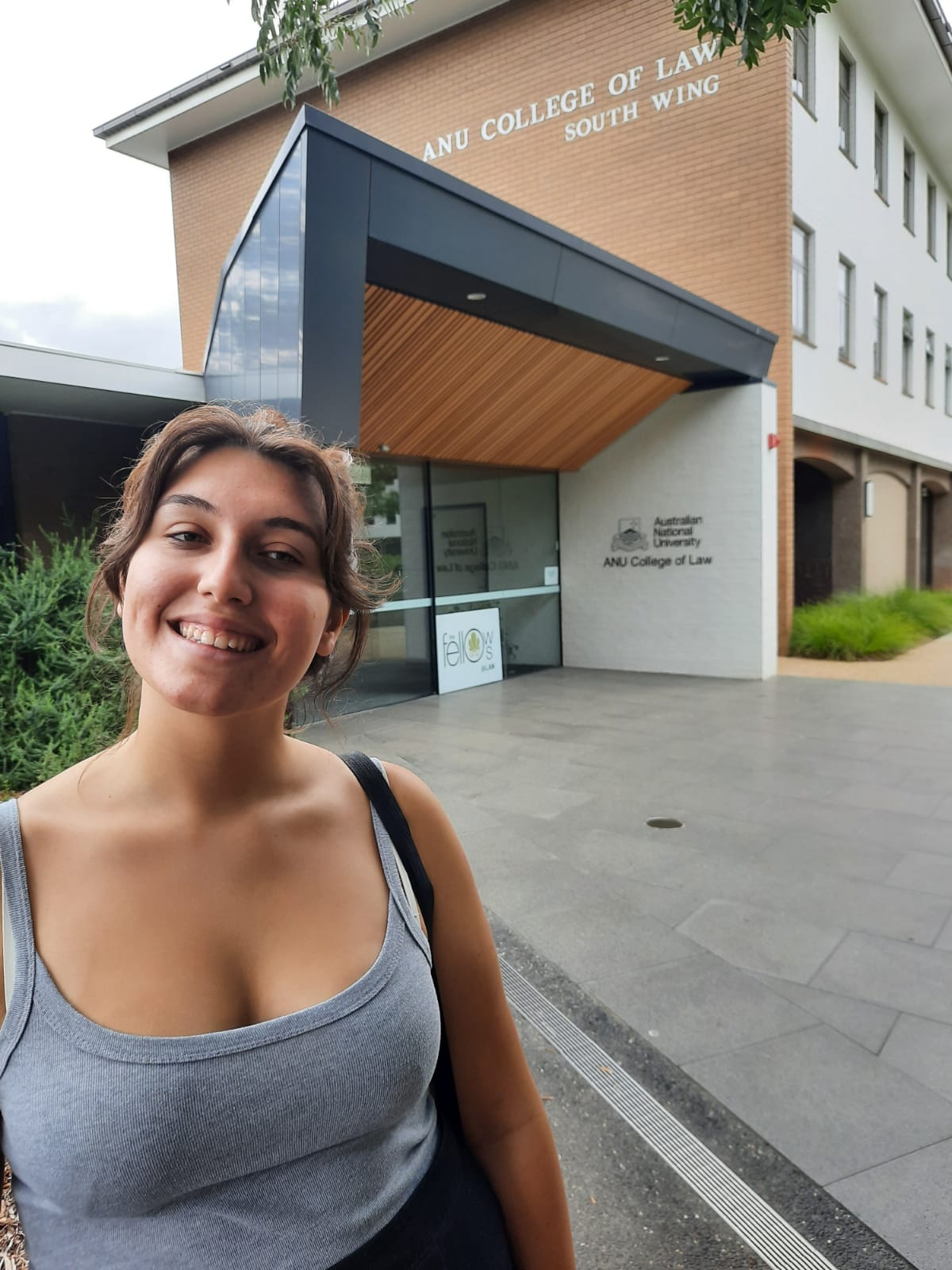 A photo of Margherita smiling in front of the ANU College of Law South Wing building. She has olive skin and brown hair in a bun, and is wearing a grey singlet. The entry to the building and some greenery is behind her.