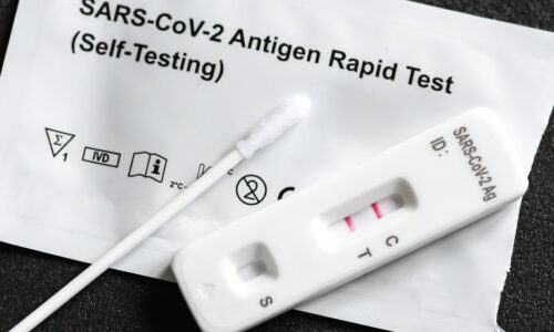 A COVID-19 rapid antigen test kit. A nasal swab and positive test cassette sit on top of the packet that has been torn open.