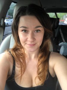A photo of Azure, a white woman, from the shoulders up, sitting in her car and smiling close-mouthed at the camera. She has long brown ombre hair loose about her shoulders and is wearing a black tank top. In the background is a baby seat for the car. 