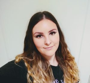 A photo of Bethany, a young white woman, from the shoulders-up standing in front of a white wall, wearing a black t-shirt and her wavy, brown ombre hair loose about her shoulders. She is smiling at the camera, close-mouthed, wearing light makeup and black, winged eyeliner.