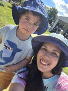 An outdoor selfie of Paloma, a 2-year-old girl with short curly brown hair wearing a blue bucket hat, blue shirt with a shark on it and orange shorts. On the right is Blanca smiling wearing a blue bucket hat, multi-colour tie dye shirt, has long dark hair and olive skin tone.