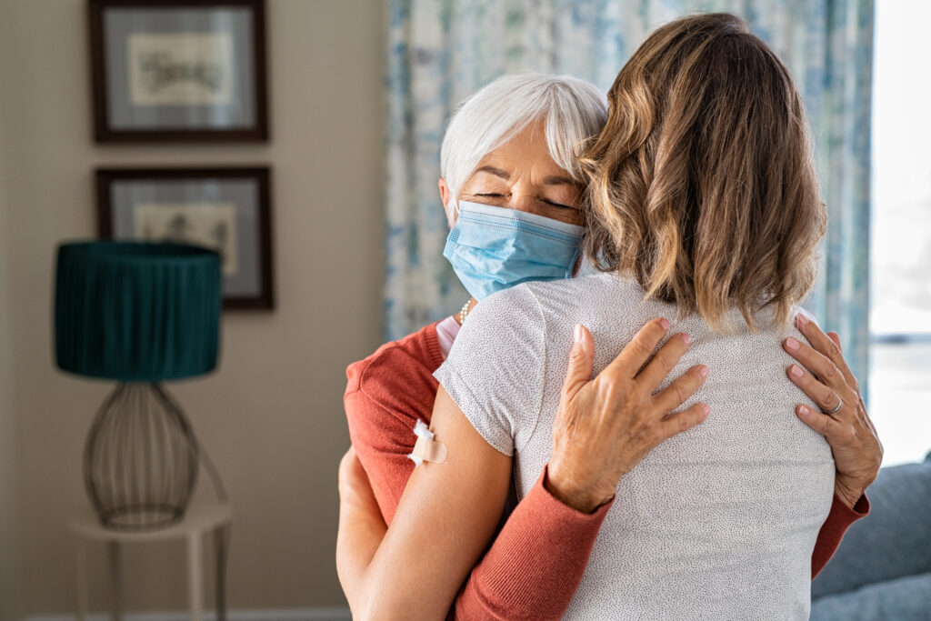 Senior woman wearing face mask hugging immunized daughter after covid-19 vaccination shot. Elderly grandmother hugging adult granddaughter after covid vaccine jab. Immunity and end of covid19 pandemic