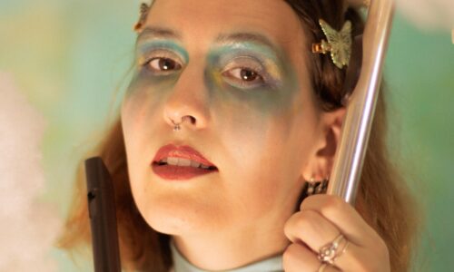 A photo of the Emerald Ruby, posing with a silver flute in one hand and a wooden recorder in the other. She has fair skin, shoulder length brown hair with butterfly clips, and is wearing pink lipstick and blue and gold eyeshadow in an angelic style. She is wearing a light blue and white turtleneck shirt, standing in front of a background of clouds and blue/green colours that had been staged in the background.