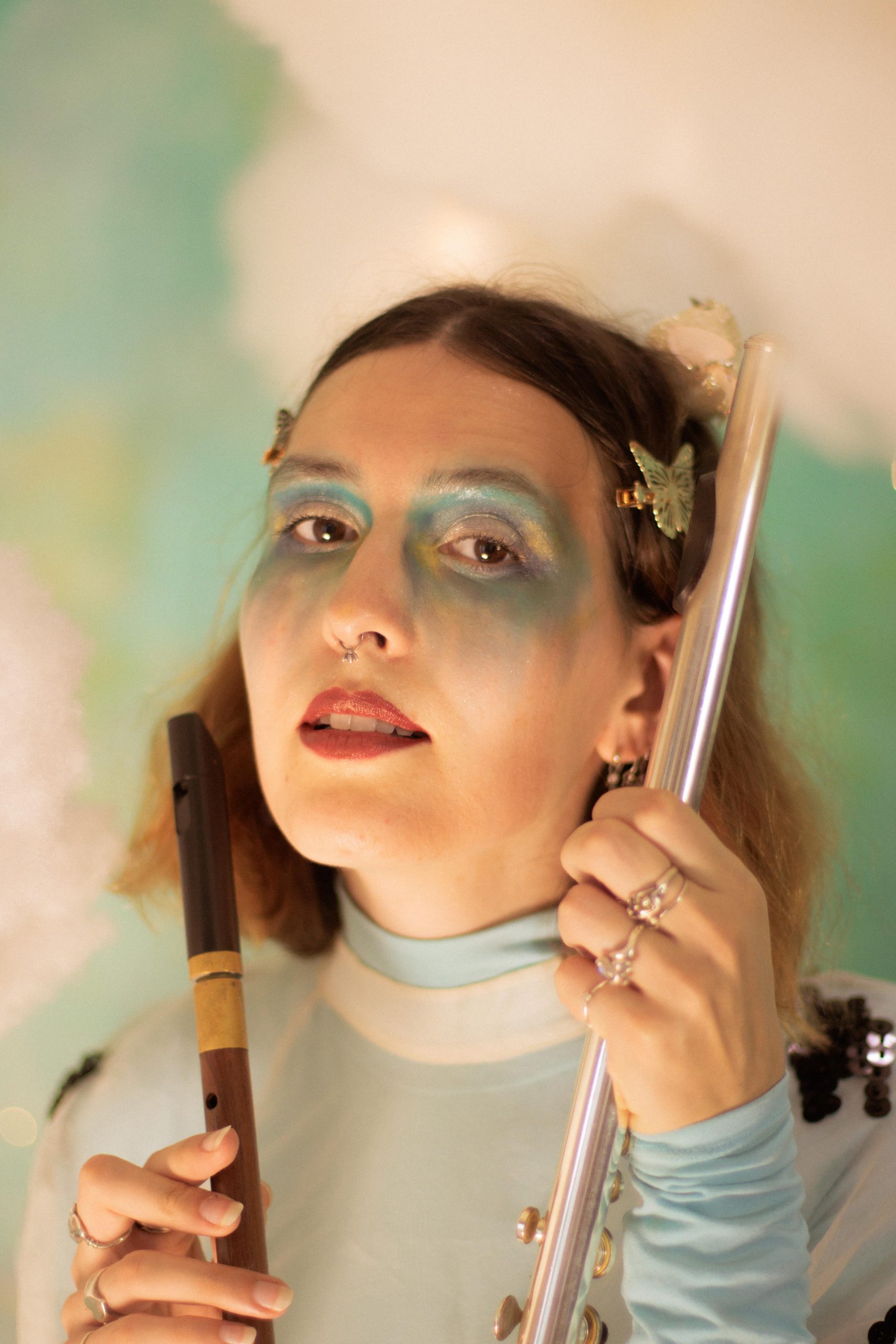 A photo of the Emerald Ruby, posing with a silver flute in one hand and a wooden recorder in the other. She has fair skin, shoulder length brown hair with butterfly clips, and is wearing pink lipstick and blue and gold eyeshadow in an angelic style. She is wearing a light blue and white turtleneck shirt, standing in front of a background of clouds and blue/green colours that had been staged in the background.