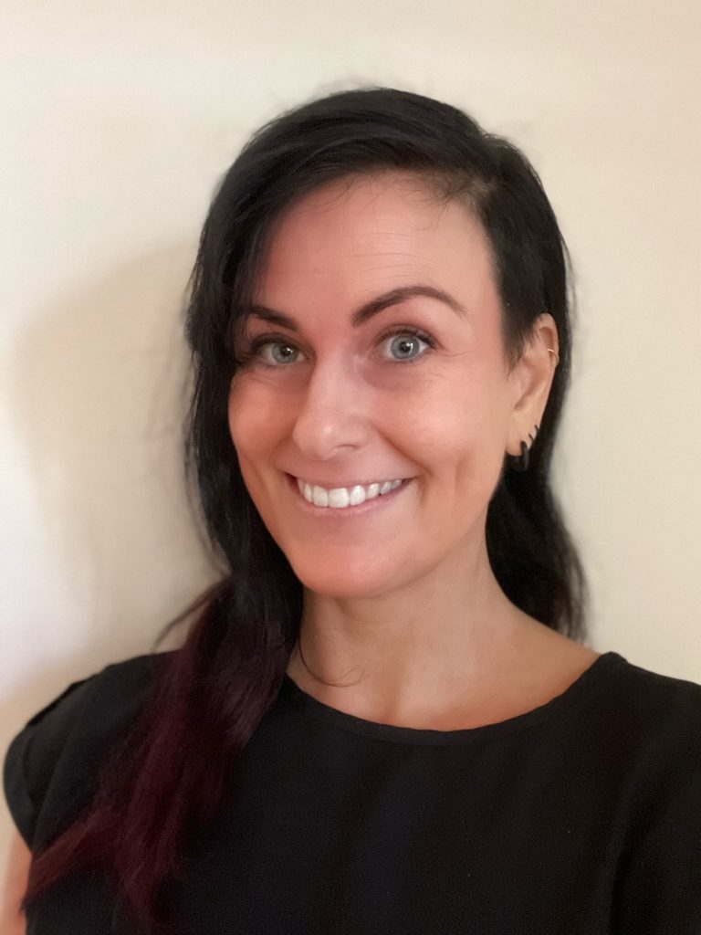 A portrait photograph of Karina. She is smiling, looking at the camera and wearing a black top. Karina has long black hair with dark red ends and one side shaved, blue eyes and multiple piercings in her ear.