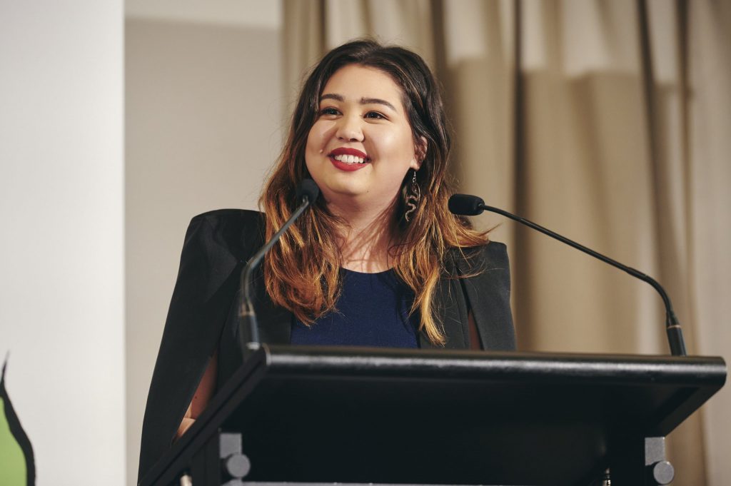 An image of Kat smiling at a black podium with two microphones. They have light brown skin and long brown and copper hair. They are wearing red lipstick, snake earrings, a navy shirt and black blazer. In the background is a white wall and cream stage curtains. 