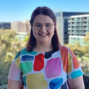 A photo of a white woman with shoulder length red-brown hair smiling directly at the camera. She is wearing light pink glasses, and her short-sleeved white top features coloured shapes. The background is blurred, somewhat showing green trees and buildings in the distance.