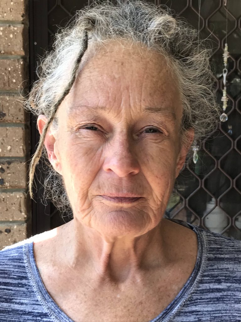 A photo of Wendy staring into the camera. She has fair skin and short curly grey hair, with one small dreadlock at the front. She is wearing a patterned blue shirt, and behind her is a brick wall and screen door.