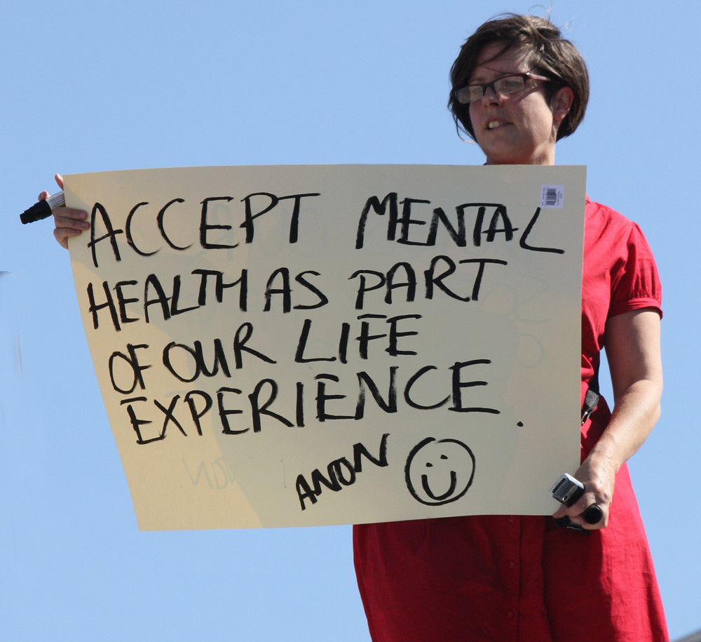 An image of a white woman with short brown hair, wearing a red dress and red glasses, holds a white protest sign with black marker that reads "Accept mental health as part of our life experience. Anon :)" In one hand the woman is holding the marker. Blue sky is visible in the background.