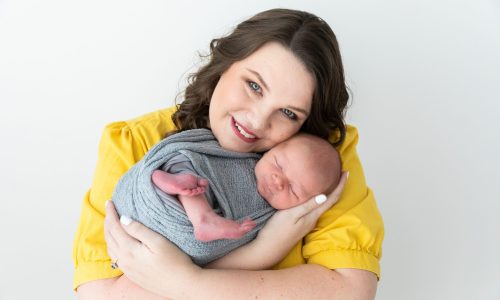 A photo of Annie smiling at the camera and cradling her newborn baby. Annie is a white woman with shoulder length brown hair wearing a bright yellow shirt. Her baby is wrapped in a blue-grey swaddle.