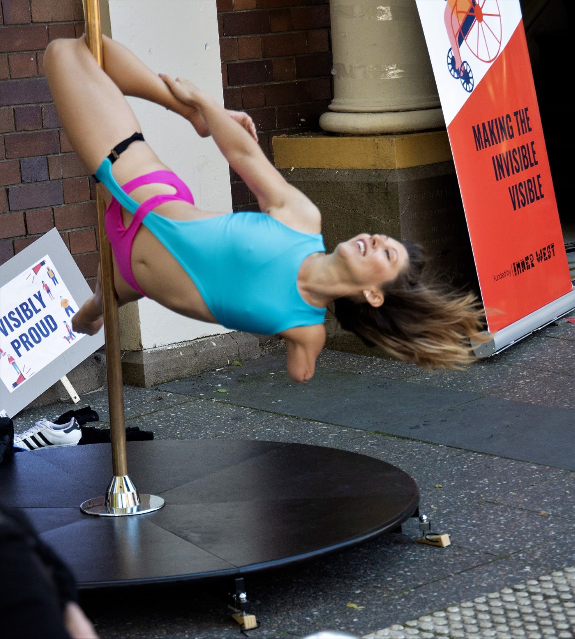 A photo of a disabled pole dancer, upside down, mid-routine on stage. She is an amputee with one arm, has long brown hair, and is wearing a blue a pink lycra body suit. Beside the stage is a pair of white and black Adidas shoes, a protest sign that reads ‘Visibly Proud’ and a red festival banner that reads ‘Making the Invisible Visible, Funded by Inner West’.