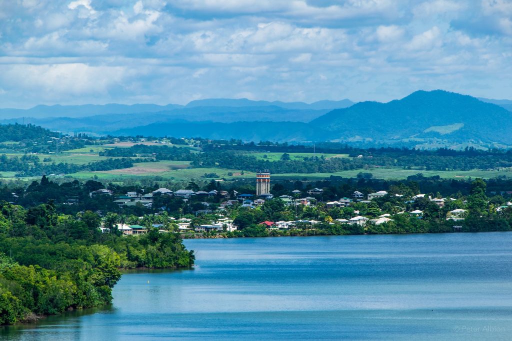 An aerial image of Innisfail in Far North Queensland, country belonging to the Mamu People. A view of the Johnstone River is visible, with trees, buildings and houses that make up the town of Innisfail pictured along the banks. In the background, green paddocks and mountains are visible, with clouds framing the image.