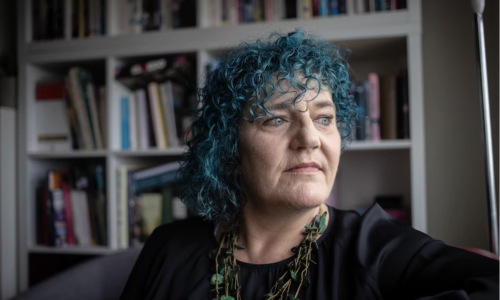 Photo of WWDA CEO Carolyn Frohmader. Carolyn has blue, short, curly hair and is looking out a window wearing a black top and green necklace in front of a book shelf.