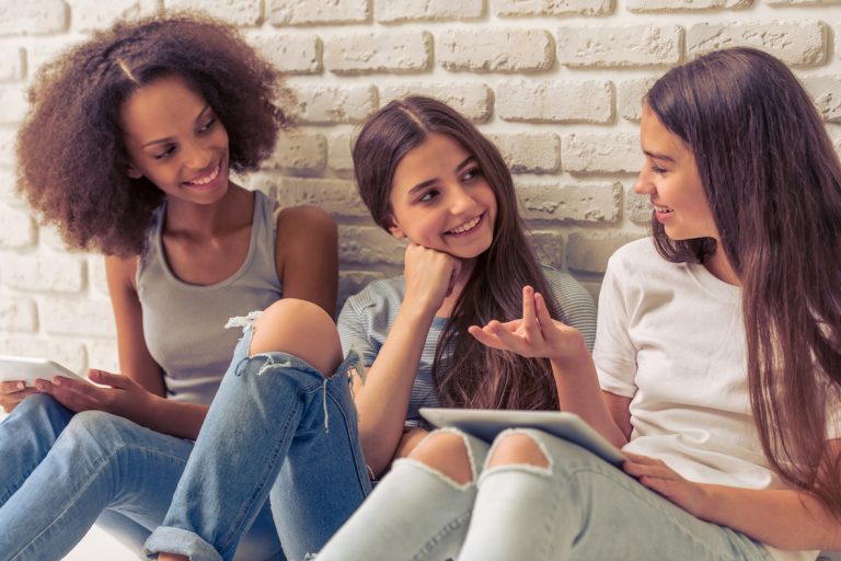 Three young women sitting against a wall talking.
