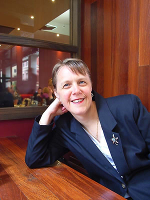 A photo of Kerryn sitting at a wooden table, leaning their head on one hand and smiling at the camera. Kerryn has white skin, short mousy brown hair, and is wearing a navy blazer and brooch over a light blue blouse.