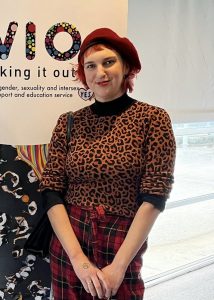 A nonbinary person with pale skin, short red hair, standing with hands in front of them smiling at the camera. 