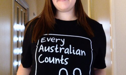 A photo of Bethany, smiling with her mouth closed and holding her black and white Every Australian Counts #DefendOurNDIS campaign shirt so it is visible. She has fair skin, long brown hair, and is standing in the hallway of a house.