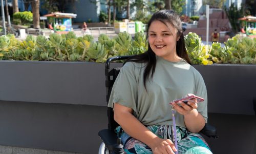 Young woman using a wheelchair smiling at the camera and holding up a smart phone. In the background is a concrete planter with green plants in it.