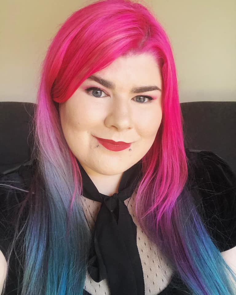 Portrait photo of Zoe, who has white skin and long hair that is a gradient of pink to blue. Zoe is wearing a black neck tie and red lipstick