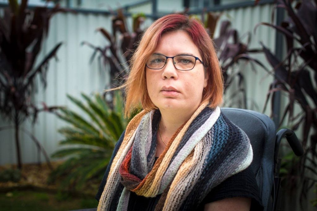 Image of Kelly Cox, Kelly has light brown hair, is wearing glasses, and a colourful scarf. There are plants that are blurry in the background.