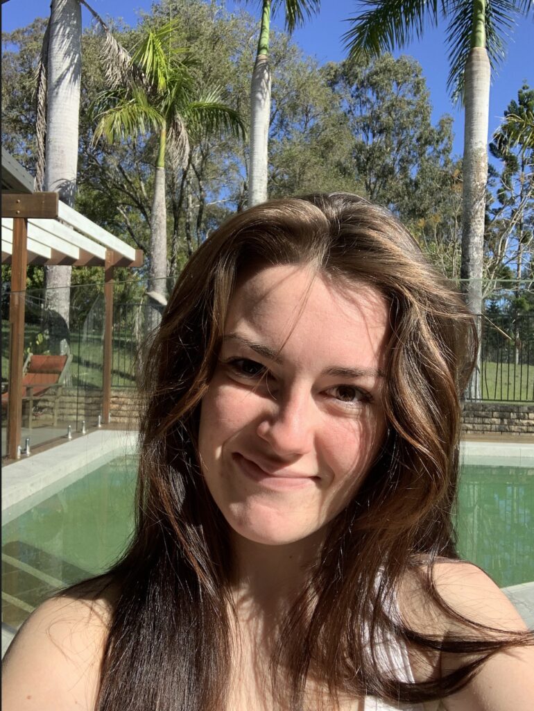 Portrait photo of Breagh smiling, outside with a pool in the background. She has white skin and very long brown hair and is wearing a white tank top.