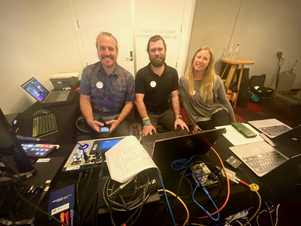 Photo of three people sitting behind a table with a lot of tech equipment. Jamie has a ponytail with a patterned top and moustache. Peter has facial hair and wearing a black top and Gabriella has long blonde hair and wearing a grey top. 