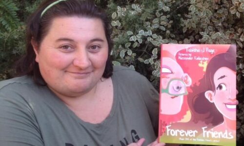 Tabitha is standing outside in front of some bushes smiling and holding her book Forever Friends. Tabitha has white skin, short brown hair held back by a thin headband and is wearing a green shirt with the word ‘daring’ visible.