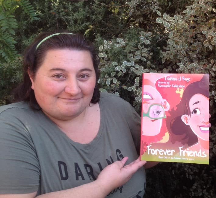 Tabitha is standing outside in front of some bushes smiling and holding her book Forever Friends. Tabitha has white skin, short brown hair held back by a thin headband and is wearing a green shirt with the word ‘daring’ visible.