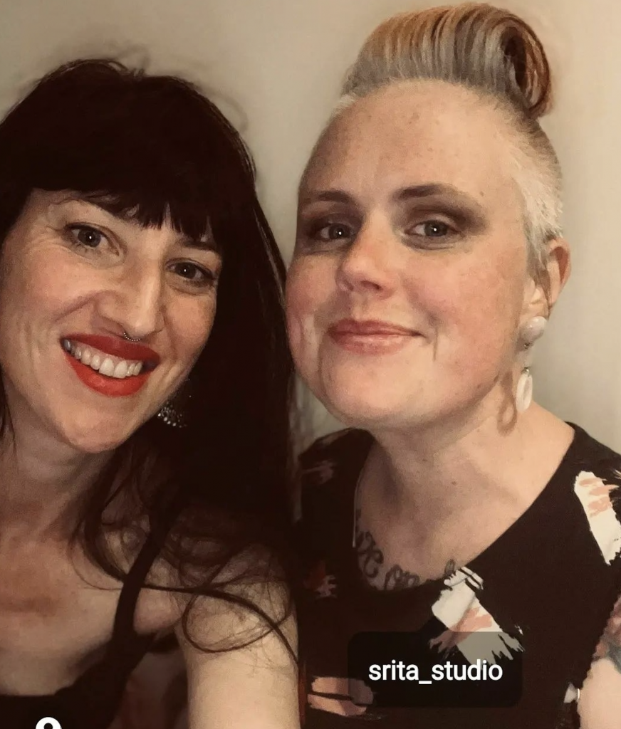 Photo of author, Sarah Langston and friend. Sarah, on the right has long dark hair with a fringe and is smiling. She is wearing bright red lipstick. Her friend, on the left has white blonde hair pushed back in a comb over. She is wearing long dangling earrings and a black top.
