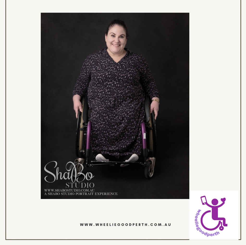 A white square with a black rim around it. In the middle is a square photo of Amber from wheeliegoodperth. She is a wheelchair user and is wearing a black dress and has dark hair that is pulled back. In the bottom left hand corner of the image is white cursive text reading: 'ShaBo Studio. www.shabostudio.com.au. A shabostudio portrait experience.' In the bottom right hand corner is a purple icon of a person using a wheelchair holding a smart phone in one hand and a heart in the other. Curved around the wheelchair is text: 'wheeliegoodperth.' At the bottom of the image is text in black: 'www.wheeliegoodperth.com.au.'