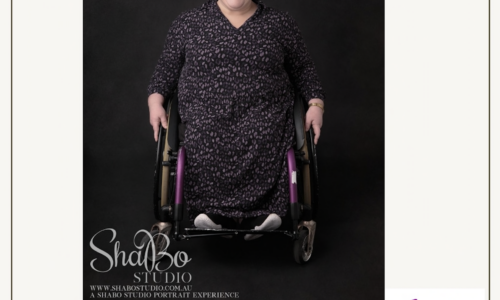 A white square with a black rim around it. In the middle is a square photo of Amber from wheeliegoodperth. She is a wheelchair user and is wearing a black dress and has dark hair that is pulled back. In the bottom left hand corner of the image is white cursive text reading: 'ShaBo Studio. www.shabostudio.com.au. A shabostudio portrait experience.' In the bottom right hand corner is a purple icon of a person using a wheelchair holding a smart phone in one hand and a heart in the other. Curved around the wheelchair is text: 'wheeliegoodperth.' At the bottom of the image is text in black: 'www.wheeliegoodperth.com.au.'