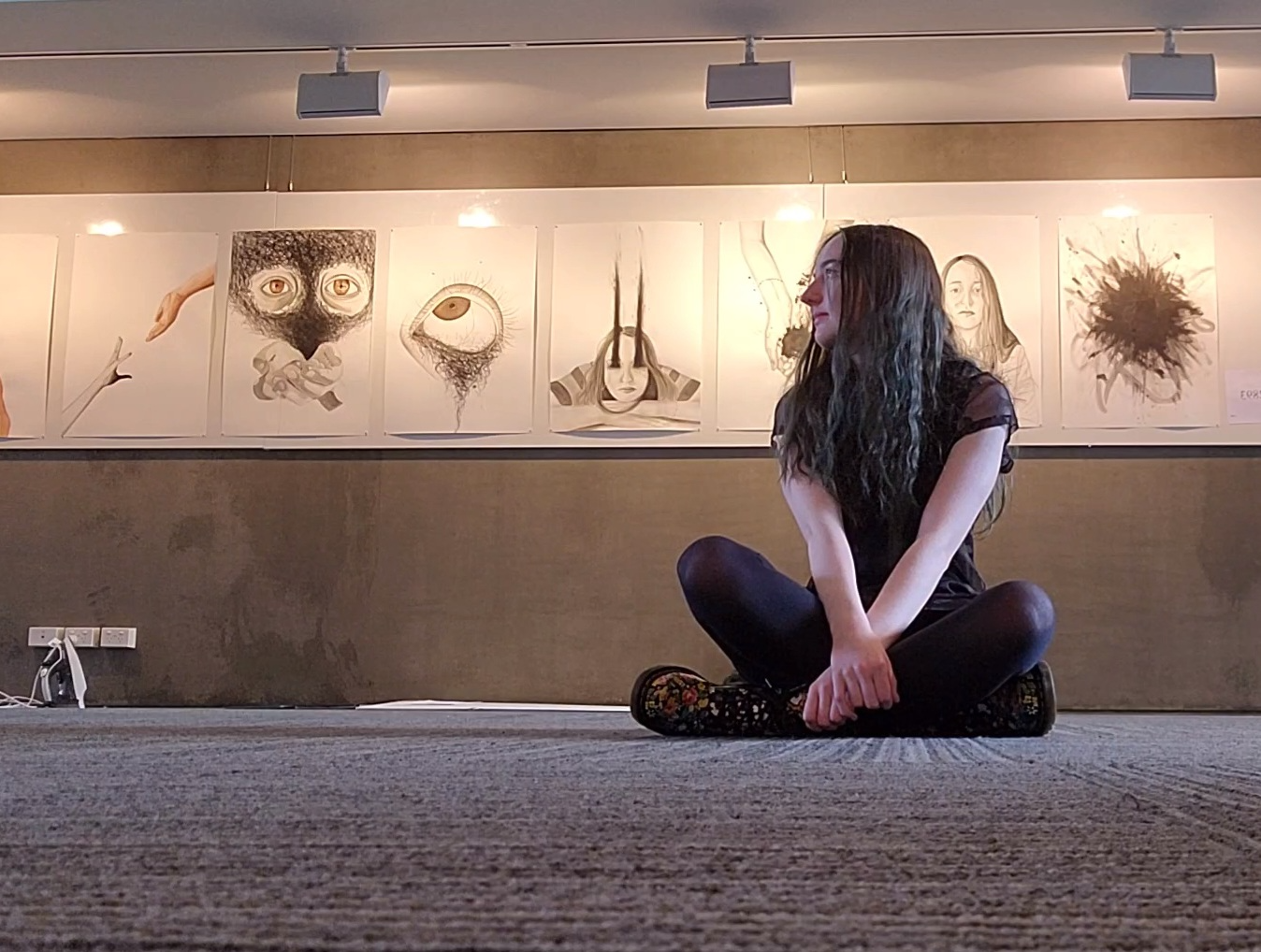 [Image: Young girl wearing all black sitting cross-legged with an exhibit of her art behind her.]