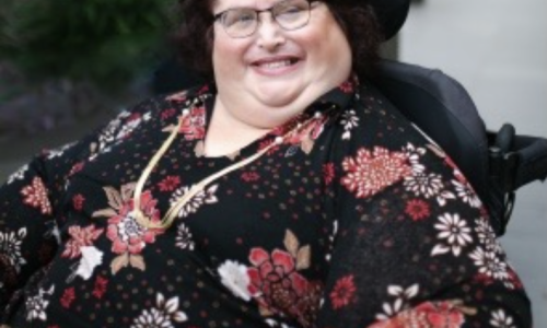 Photo of Karin Swift who has short black/ red hair and is wearing glasses with a black red floral top on. She is sitting in her wheelchair which trees in the background.