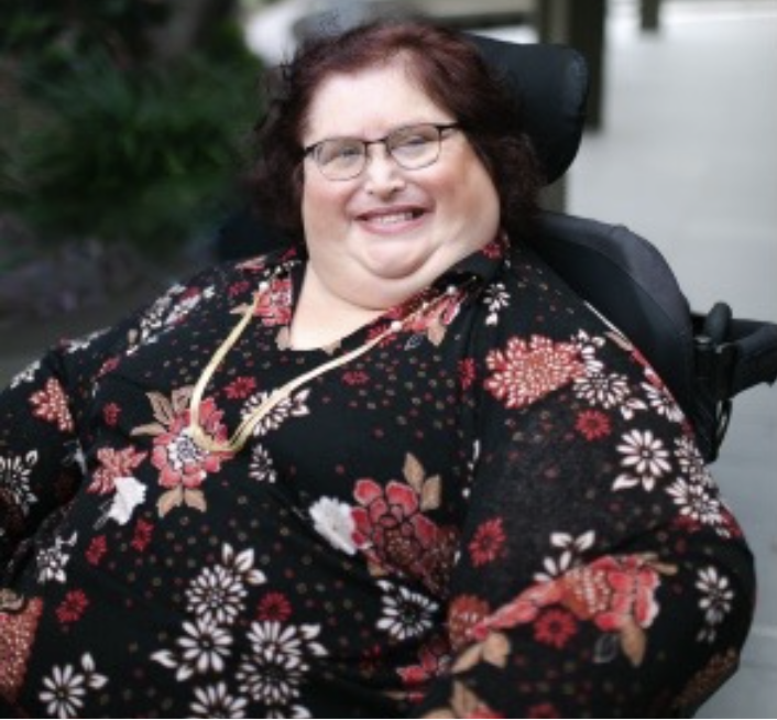 Photo of Karin Swift who has short black/ red hair and is wearing glasses with a black red floral top on. She is sitting in her wheelchair which trees in the background.