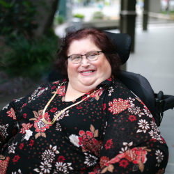 Photo of Karin Swift who has short black/ red hair and is wearing glasses with a black red floral top on. She is sitting in her wheelchair which trees in the background.
