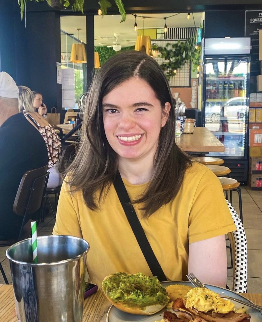 A photo of Nicola, a woman in her twenties, smiling with medium length brown hair and brown eyes. Nicola is sitting in a café, wearing a mustard yellow t-shirt and there is a black strap of a cross-body shoulder bag across her chest. In front of her is a café table with a plate of breakfast and a large metal cup, and behind her are other tables and people in the café, hanging lights and plants.  