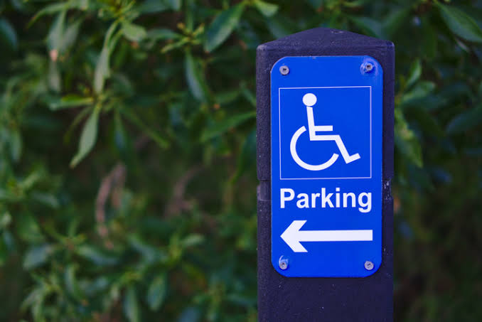 A photo of a blue and white disability parking sign. The background of the sign is blue. There is a white wheelchair symbol with the word Parking underneath. Below that is a white arrow pointing to the left.