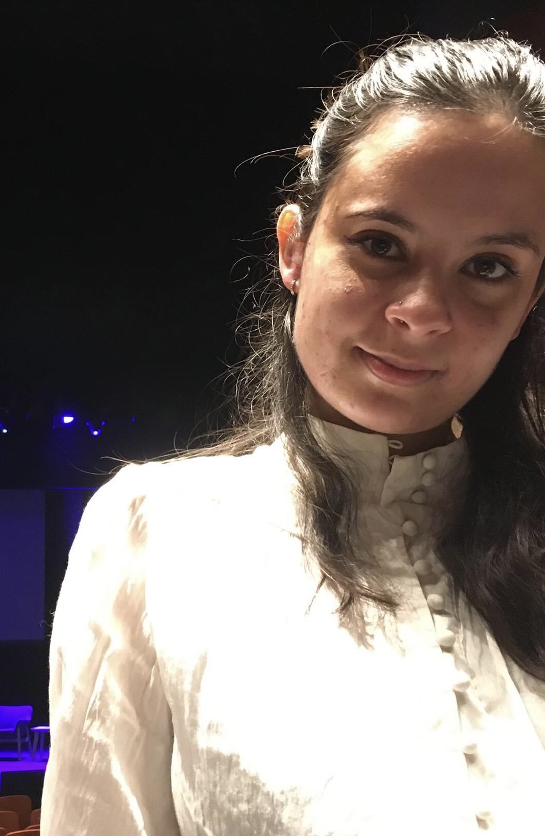 A mixed-race, medium-skinned woman in her mid-twenties. She has dark brown hair in a half-up half-down style, and is wearing a white button-up shirt.