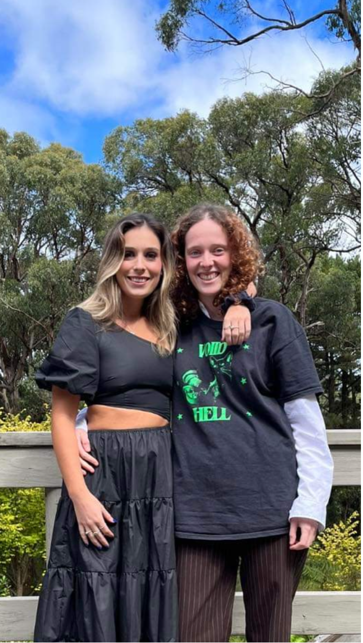 [Image description: Two feminine featured people are looking at the camera smiling. My sister, on the left, has her arm around my shoulders. She’s wearing a black crop top and a long black flowing skirt. I’m wearing a black and green t-shirt and brown pin-striped pants. Behind us are tall gum trees.]