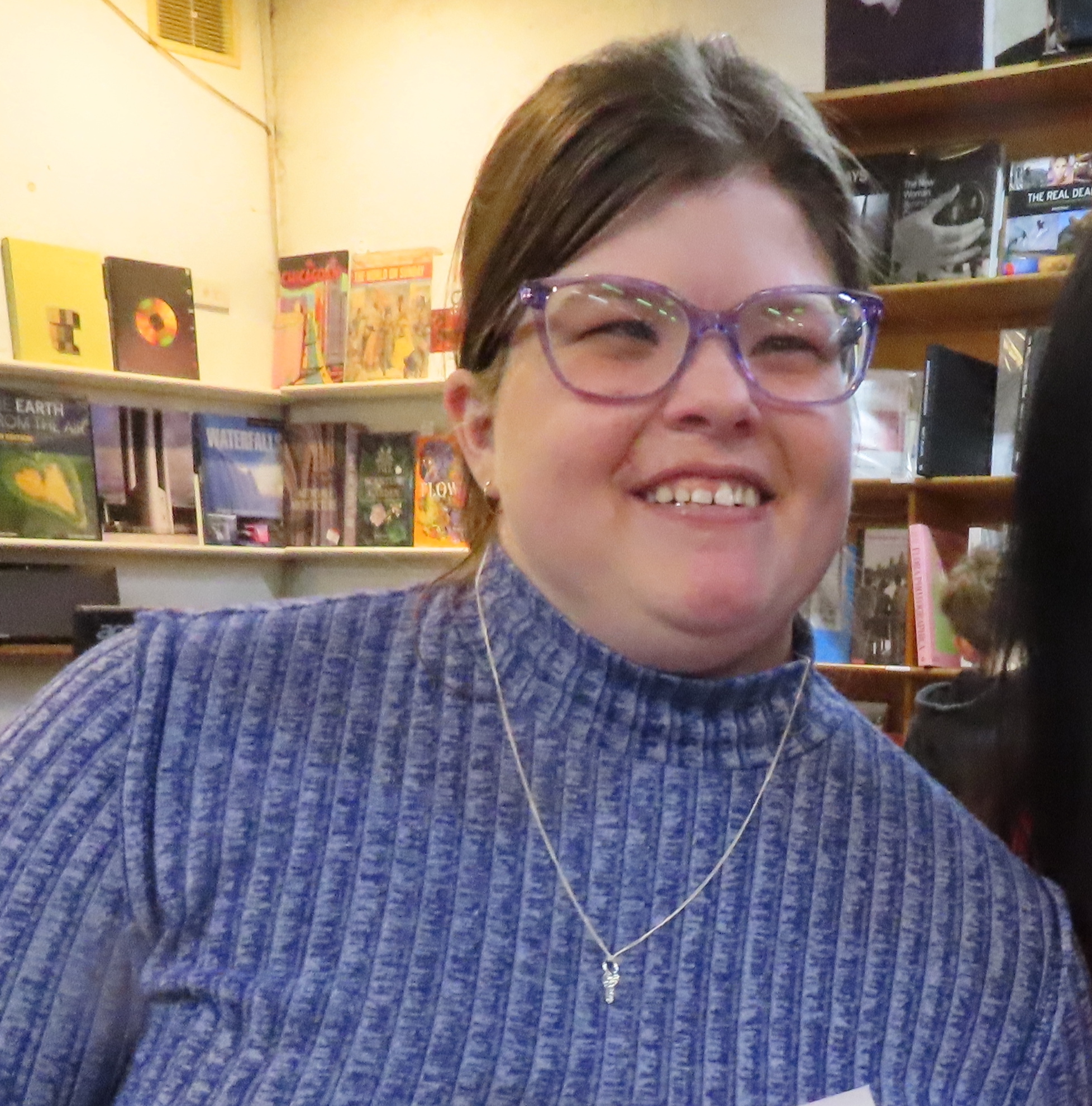 Image of Ashleigh with purple glasses standing in a bookshop. She has a long sleeved blue top on and has brown hair, and she is white.