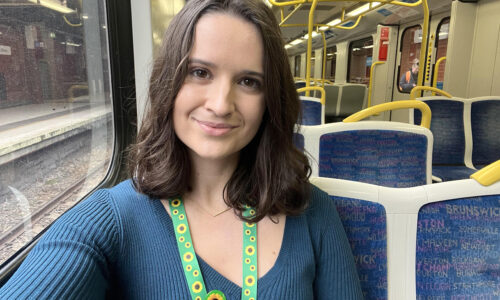 A young woman with short brown hair is sitting inside a train with yellow and blue seats in the background. She is smiling and wearing a bright green and yellow hidden disability sunflower lanyard and badge.
