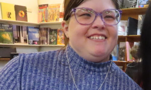 Image of Ashleigh with purple glasses standing in a bookshop. She has a long sleeved blue top on and has brown hair, and she is white.