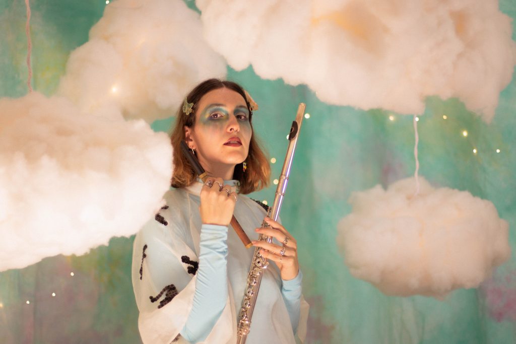A photo of the Emerald Ruby from side-on, showing her posing with a silver flute in one hand and a wooden recorder in the other. She has fair skin, shoulder length brown hair with butterfly clips, and is wearing pink lipstick, blue and gold eyeshadow in an angelic style, and multiple rings. She is wearing a light blue and white turtleneck shirt with dark sequins and is standing in front of a background of clouds, fairy lights and blue/green colours that had been staged in the background. 