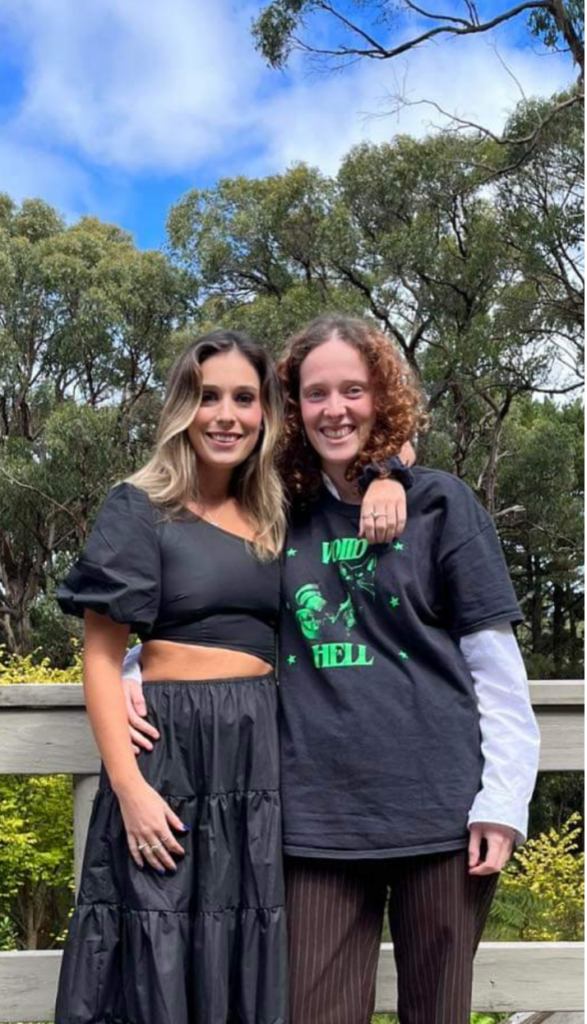Two feminine featured people are looking at the camera smiling. My sister, on the left, has her arm around my shoulders. She’s wearing a black crop top and a long black flowing skirt. I’m wearing a black and green t-shirt and brown pin-striped pants. Behind us are tall gum trees