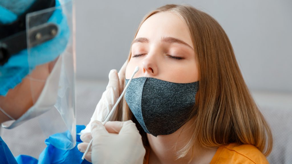 A medical professional in PPE, including gloves, a face shield and a face mask, is taking a nasal swab of a woman as part of a PCR test. The woman has her eyes closed and mask pulled down over her chin. She has fair skin and brown hair. 