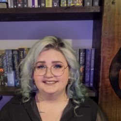 [Image: Annette is standing in front of a bookcase. Annette has green-blue hair and is wearing glasses.]
