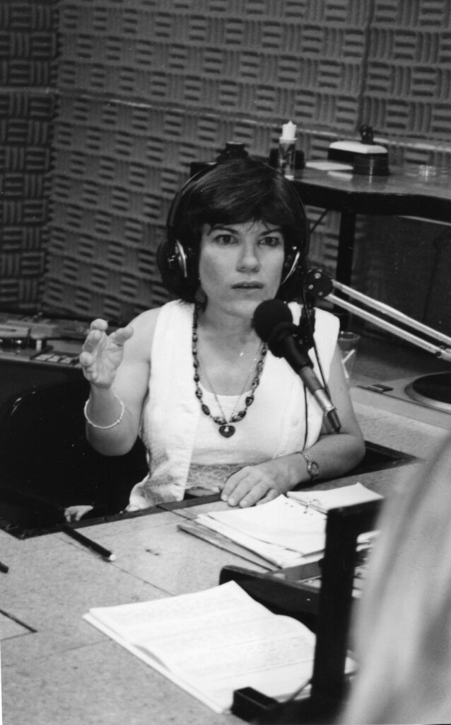 A black and white image of Suchita, a small-statured white woman with short brown hair, wearing a white shirt, chunky necklace, and black headphones. She is in a radio studio speaking into a microphone.