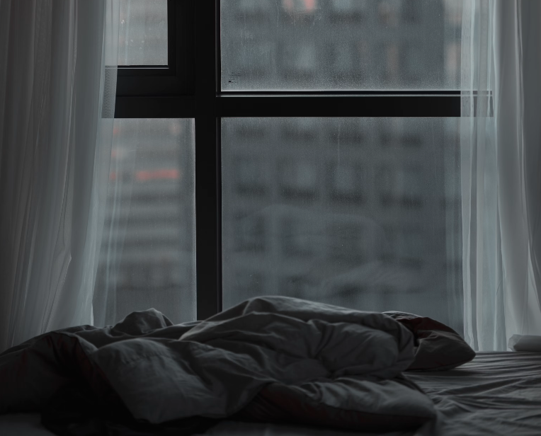 This is a grey image, with a crumpled bed sheet in the foreground. Behind this is a window with white curtains, which looks out to a grey apartment building in the distance.